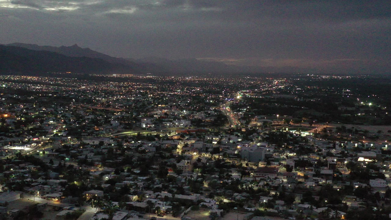 Wide aerial view of Los Cabos, Mexico at night, with city lights spread below a cloudy sky, dark mountains on the horizon, and no visible people.