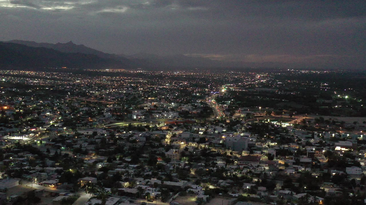 Los Cabos city lights fill the lower frame beneath a dark gray cloud deck, with mountain silhouettes left of center and no visible people.