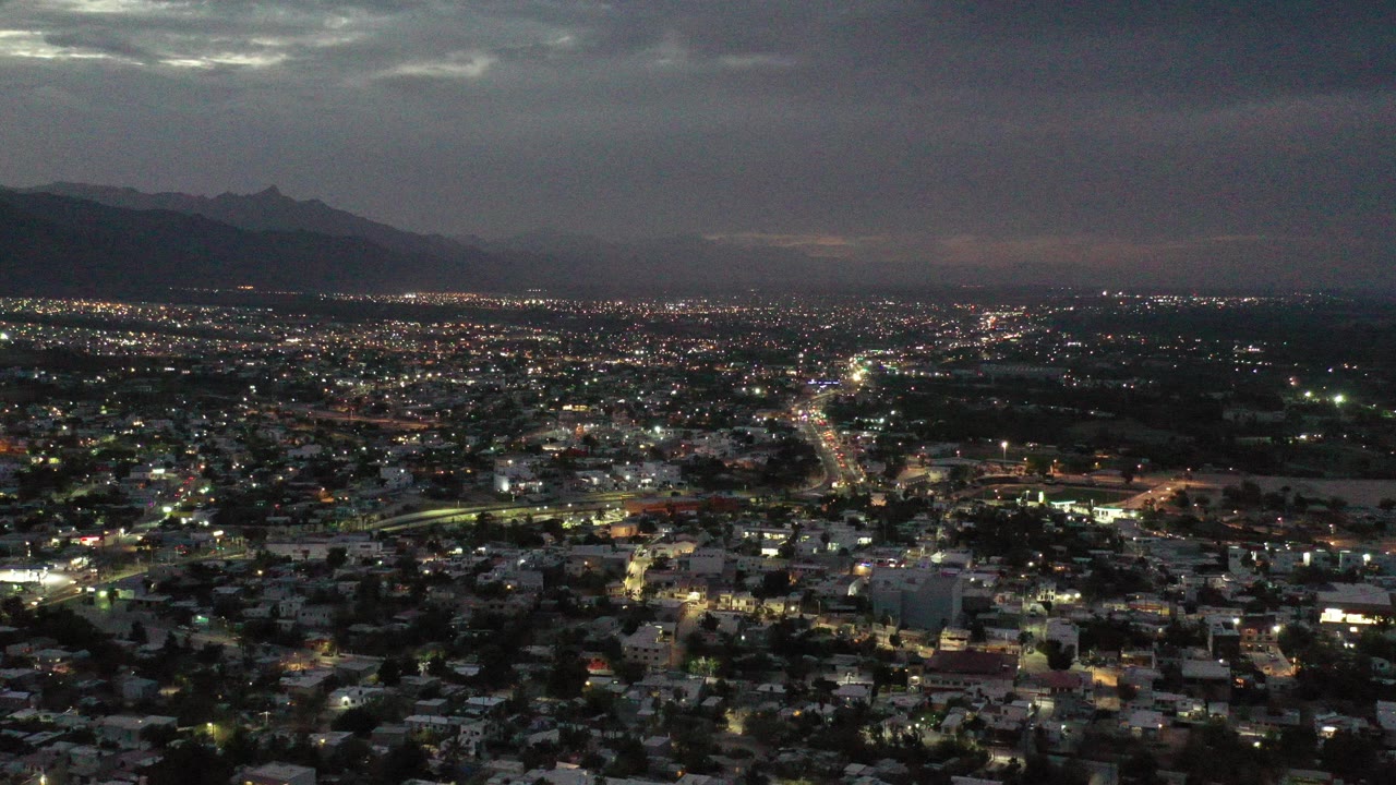 Opening frame of Los Cabos, Mexico at night, with dense city lights, a dark mountain ridge on the left horizon, heavy cloud cover, and no visible people.