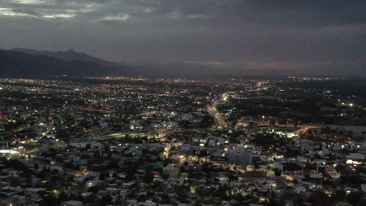 Los Cabos, Mexico appears as a wide illuminated city grid at night, with a brighter roadway near center frame, shadowed mountains in the distance, and no visible people.