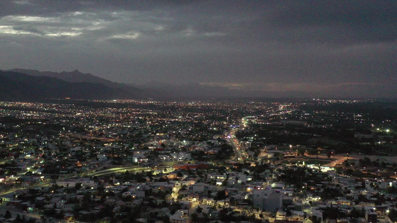 Los Cabos city lights spread below a dark evening sky, with mountain silhouettes on the left horizon and no visible people.