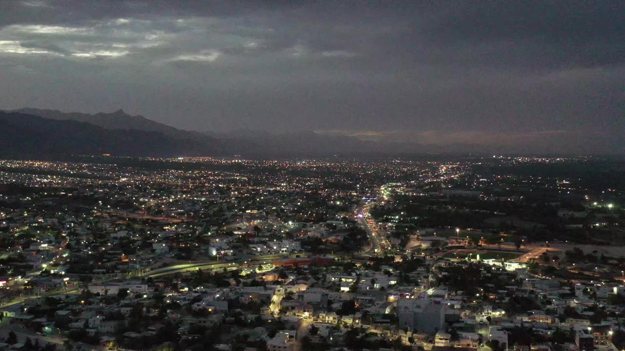 A broad Los Cabos urban grid glows under layered clouds, with the mountain ridge dimly visible across the background and no visible people.