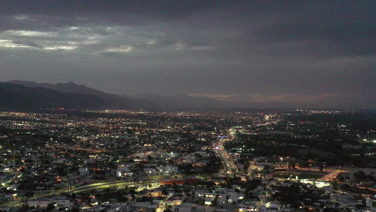 Early frame of Los Cabos, Mexico showing a wide aerial view of illuminated residential streets, low buildings, mountains on the horizon, heavy cloud cover, and no visible people.