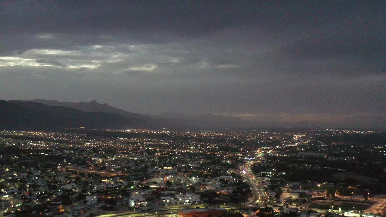 Los Cabos aerial frame showing a lit road grid across the city, shadowed mountains along the horizon, a bright opening in the gray sky, and no visible people.