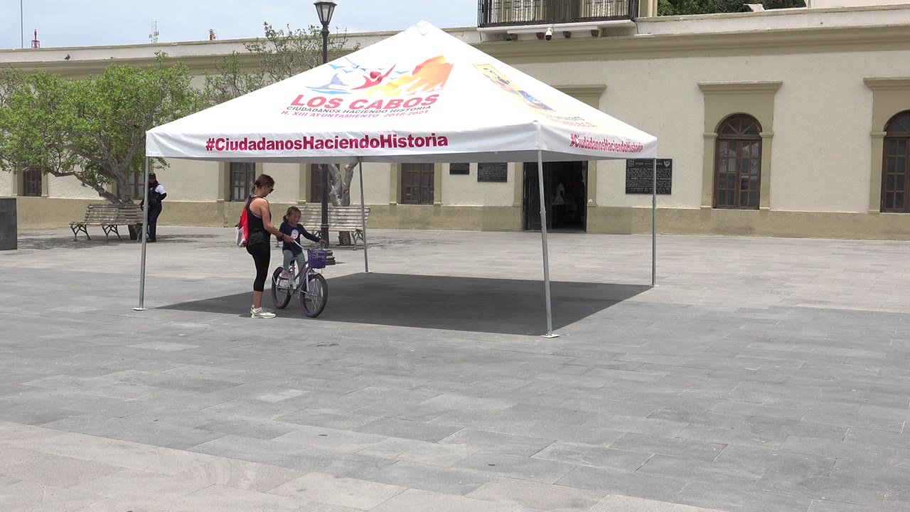 At the start of the Los Cabos, Mexico clip, a woman stands beside a child seated on a purple bicycle under a white canopy in a sunlit paved plaza.