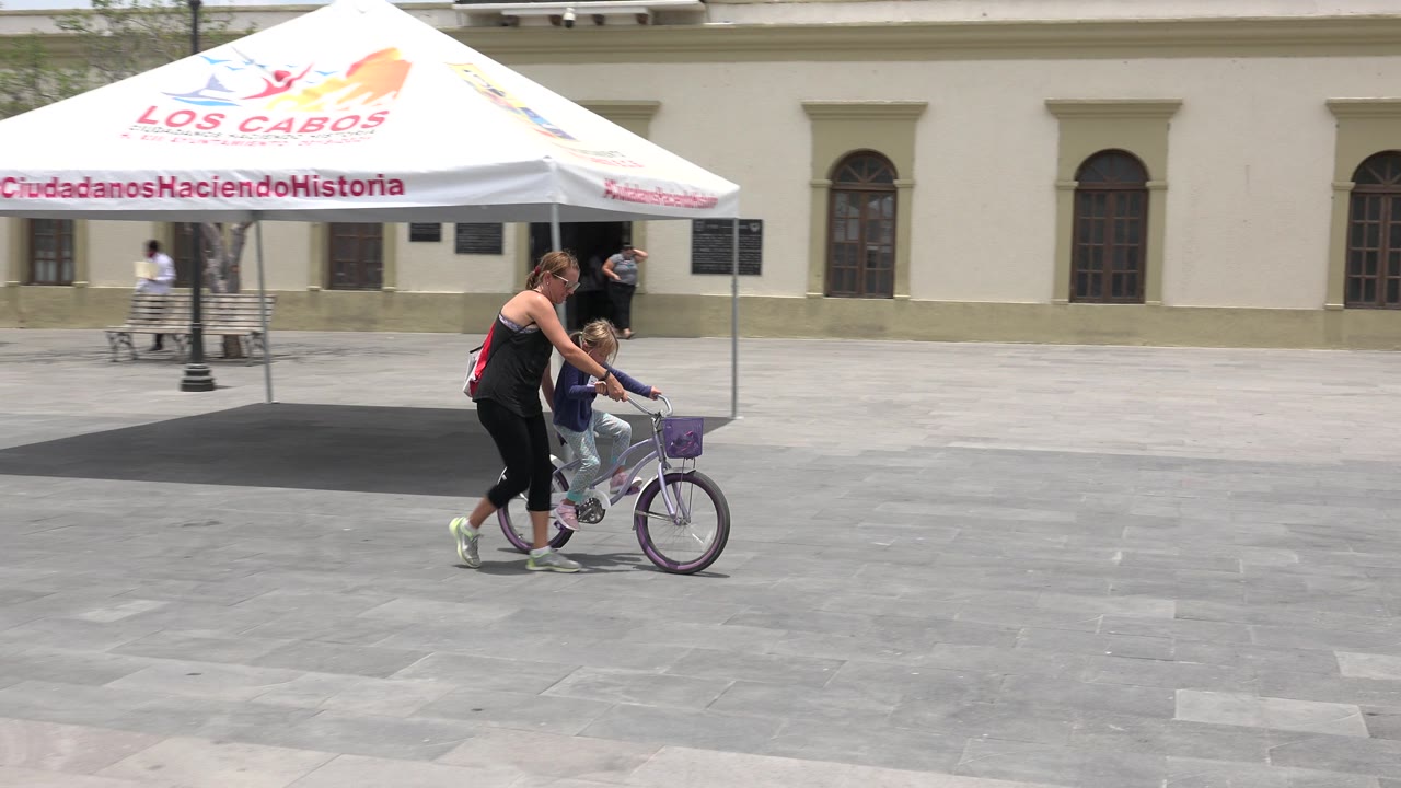 Woman helping a child ride a small purple bicycle across a paved plaza.