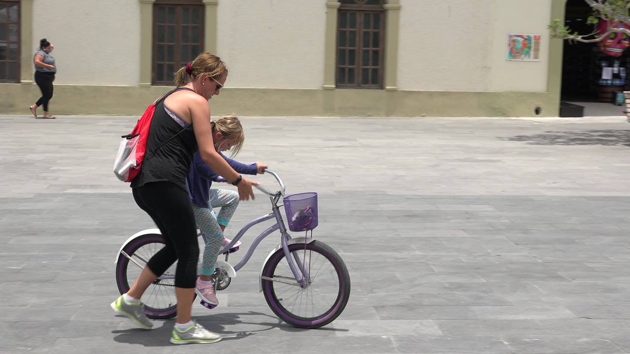 In Los Cabos, Mexico, a woman leans over a young girl on a lavender bicycle as they begin moving across a bright cement plaza.