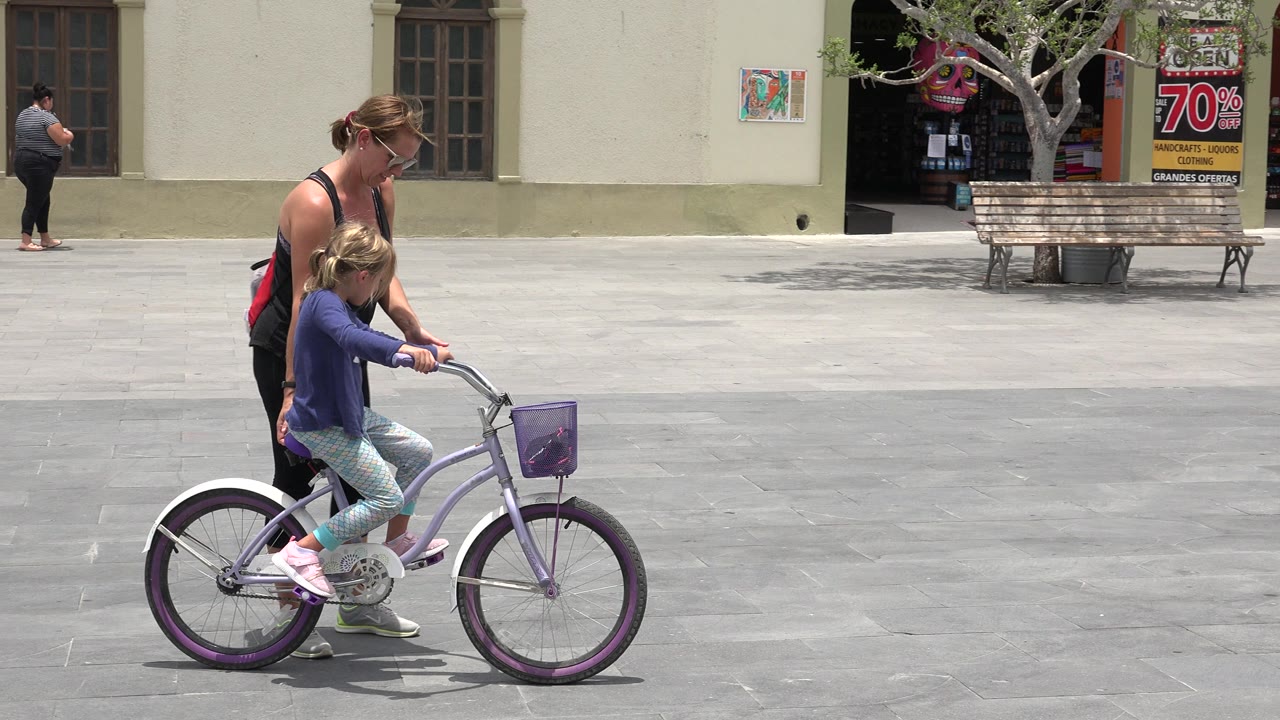 At the start of the Los Cabos clip, a young girl rides a purple bicycle while an adult woman follows closely beside her on a broad paved plaza.