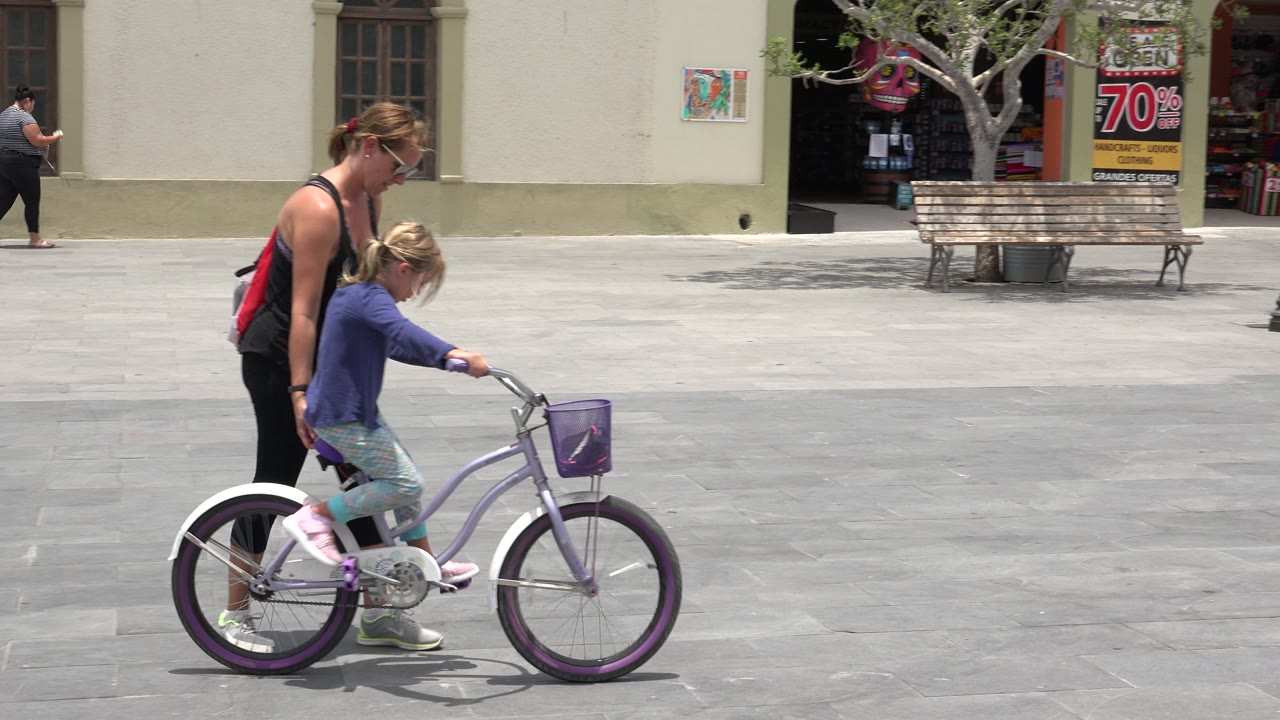Two seconds in, the girl and adult have moved farther right across the plaza, passing a bench and shaded shop entrance in the Los Cabos setting.