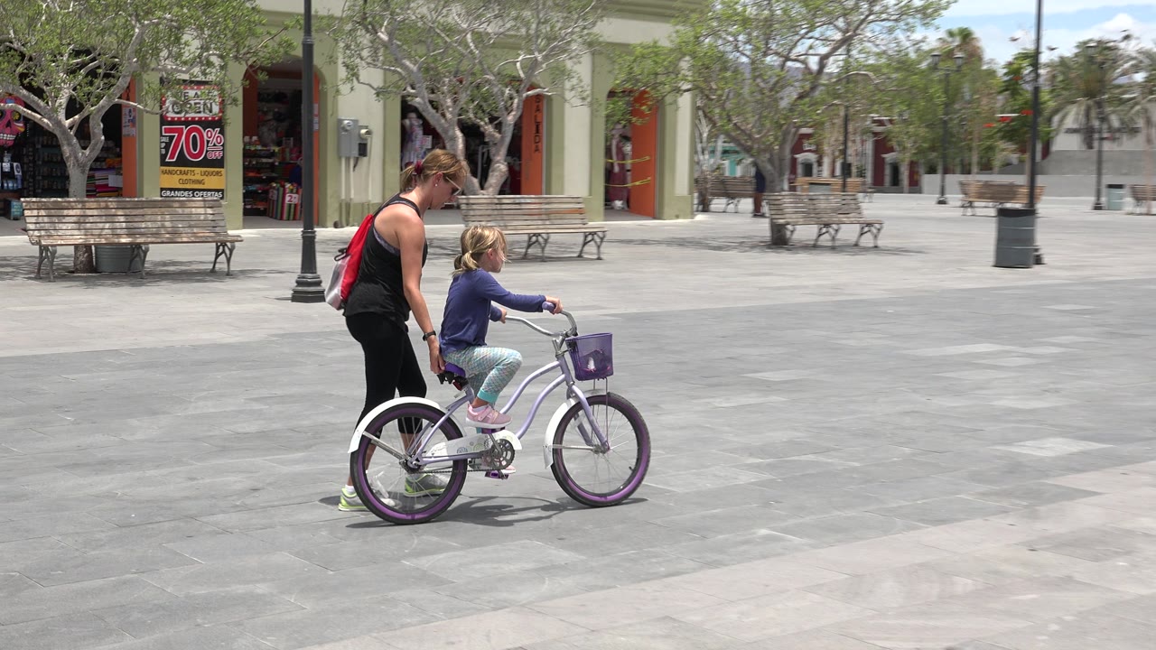 Woman walking beside a child on a bicycle in a paved plaza.