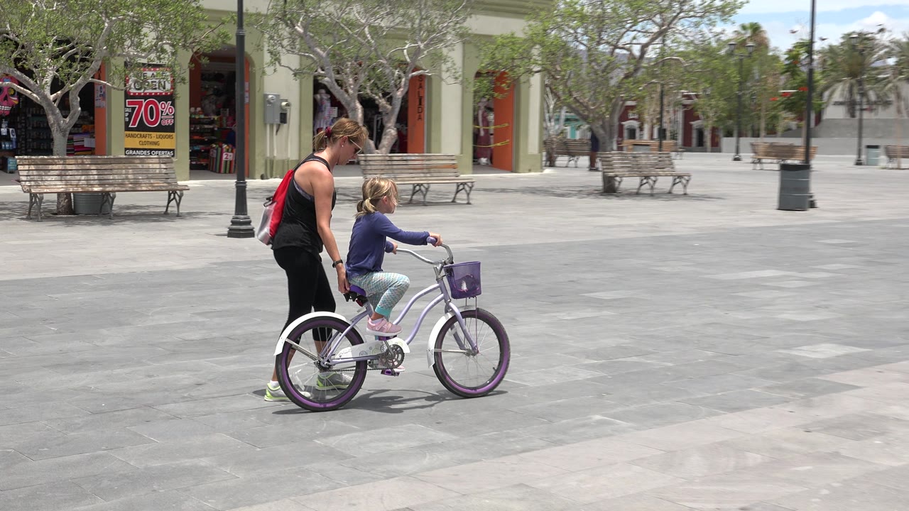The woman and child continue through the Los Cabos plaza, with the bicycle angled to the right and a shop entrance, benches, trees, and bright pavement visible.