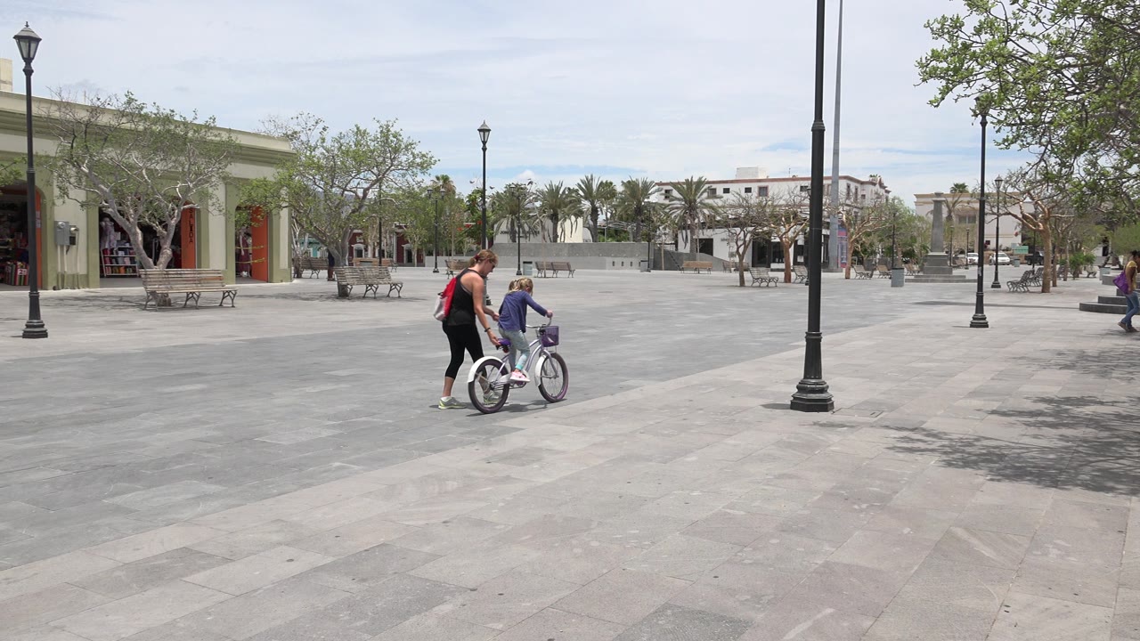 Woman guiding a child on a bicycle in a paved plaza.