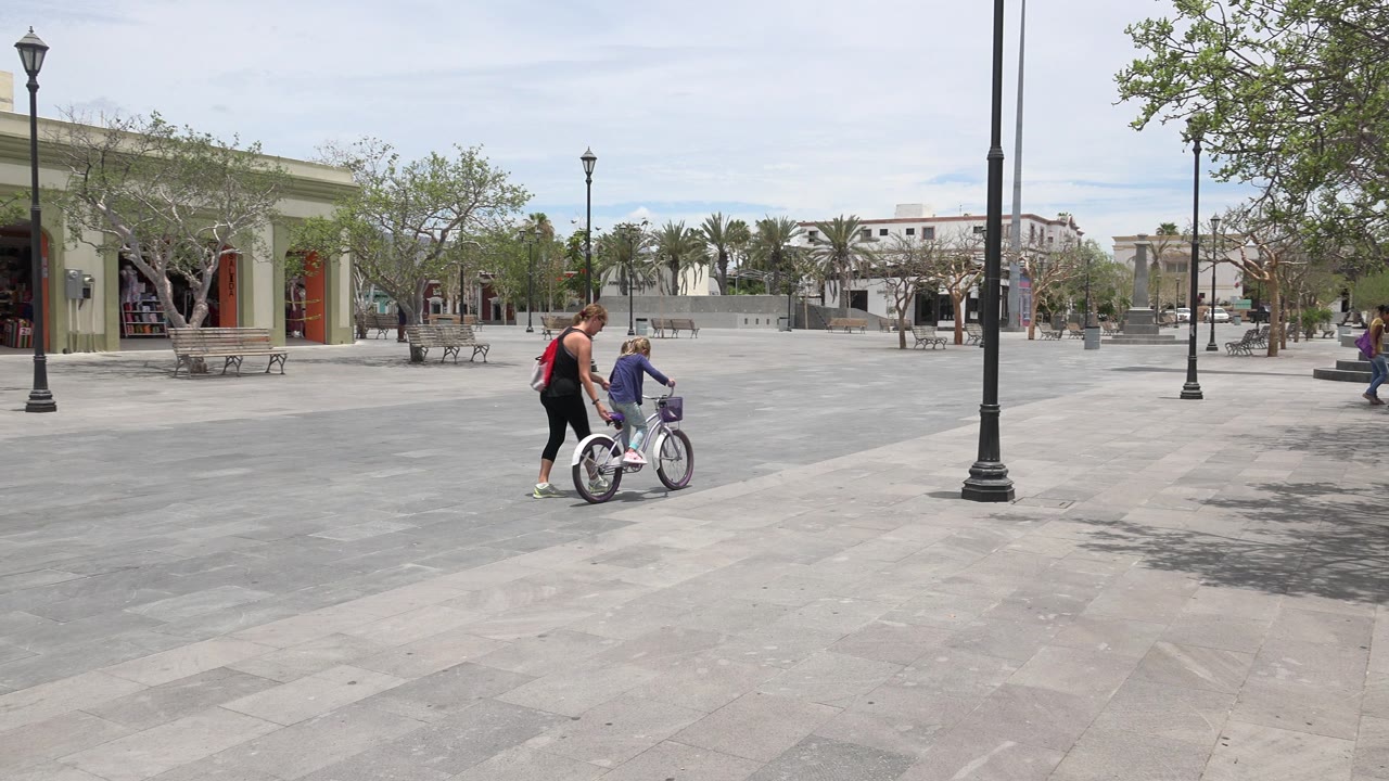The woman remains close to the child on the bicycle as they move through the open plaza, with shaded benches, lampposts, and low buildings visible in the background.