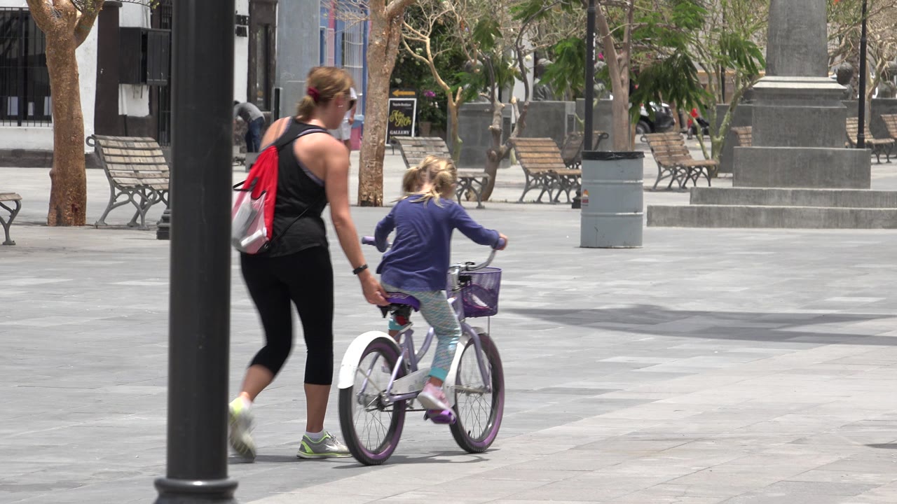 In Los Cabos, Mexico, a woman in black activewear holds a child's hand as the child sits on a small white bicycle near a dark foreground lamp post.
