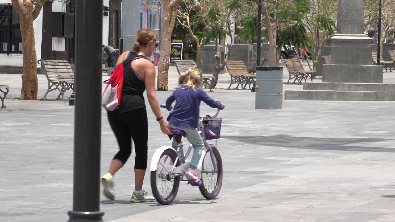 In Los Cabos, Mexico, the woman walks close behind the child on the bicycle while benches and a large stone monument remain visible across the bright plaza.