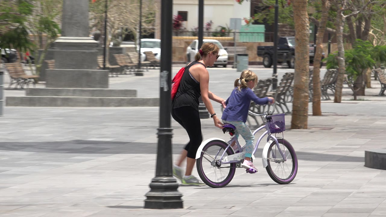 Mother walks beside daughter riding a small bicycle in a paved plaza.