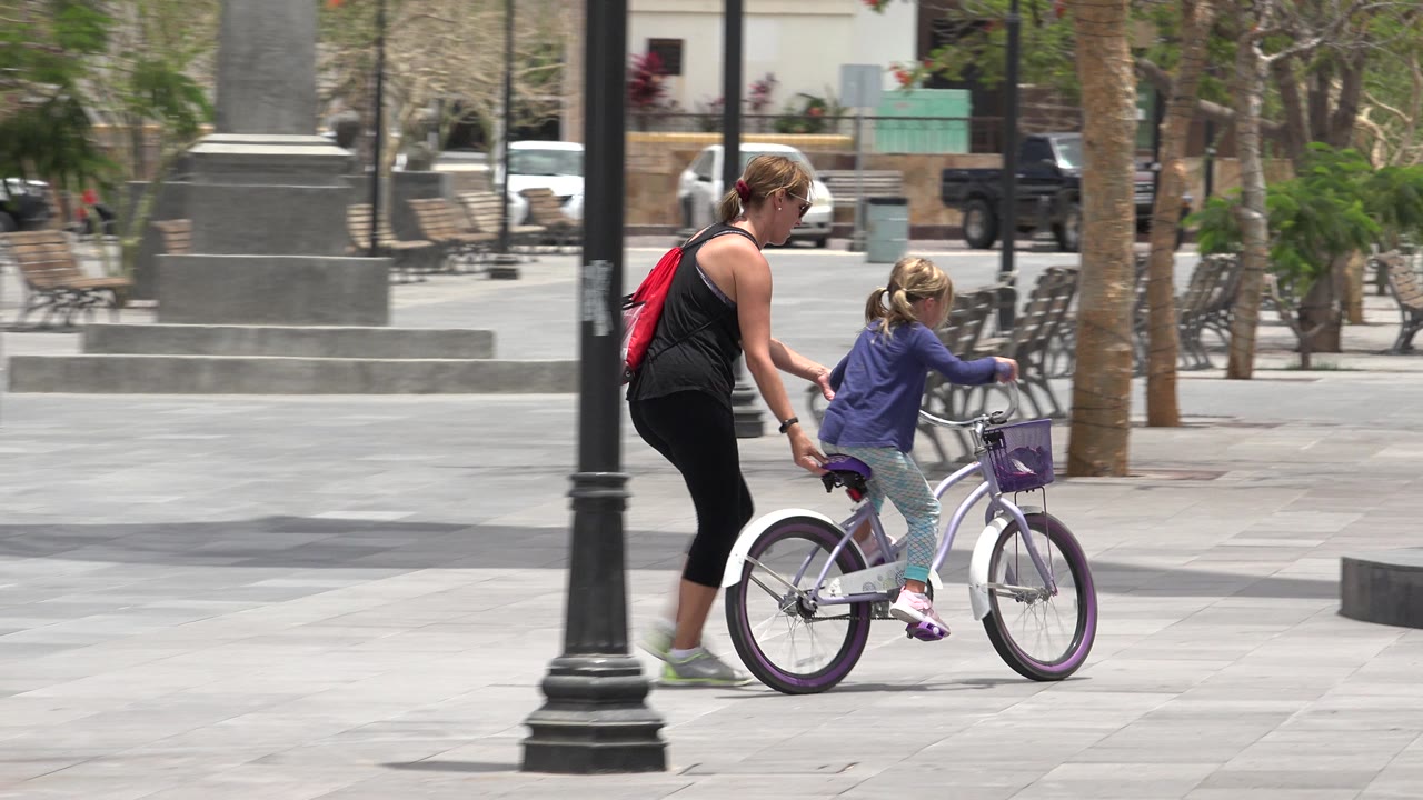 The mother keeps one hand near the child's small bicycle while they move past a dark lamp post in the Los Cabos plaza, with benches and trees behind them.