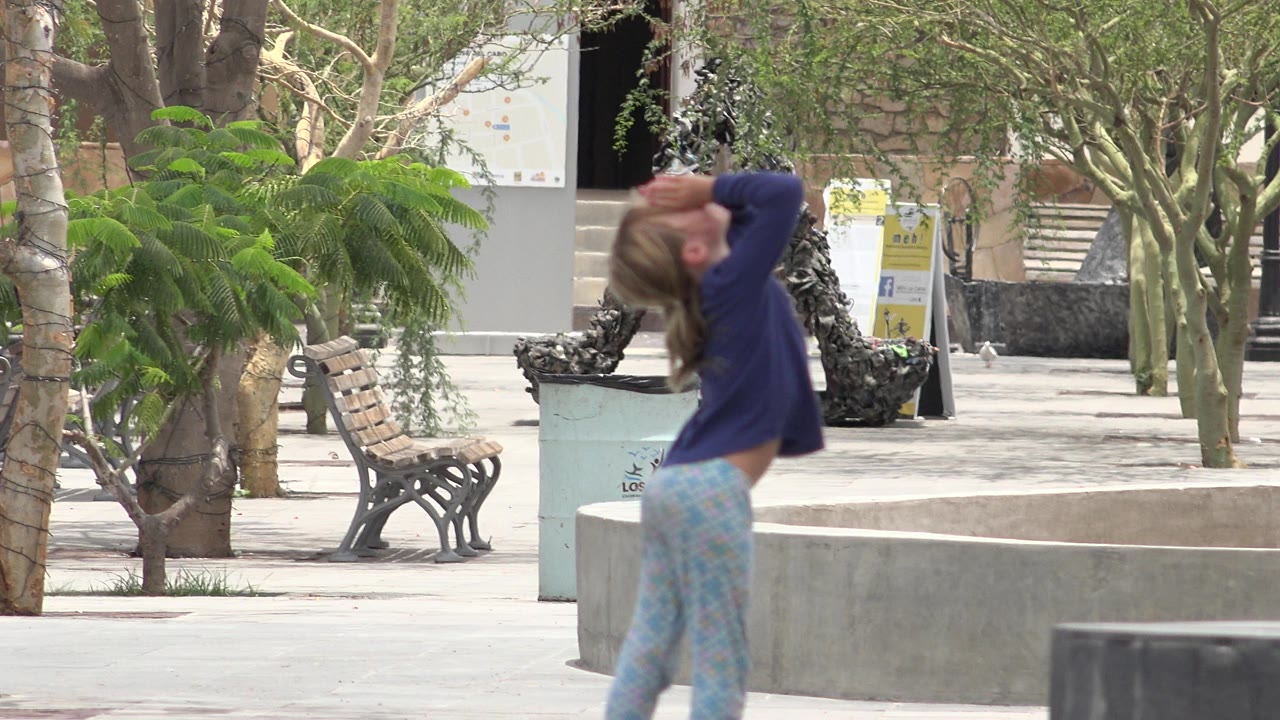 A young girl in a purple top stands in a Los Cabos, Mexico plaza with her hands near her eyes, benches and trees visible behind her.