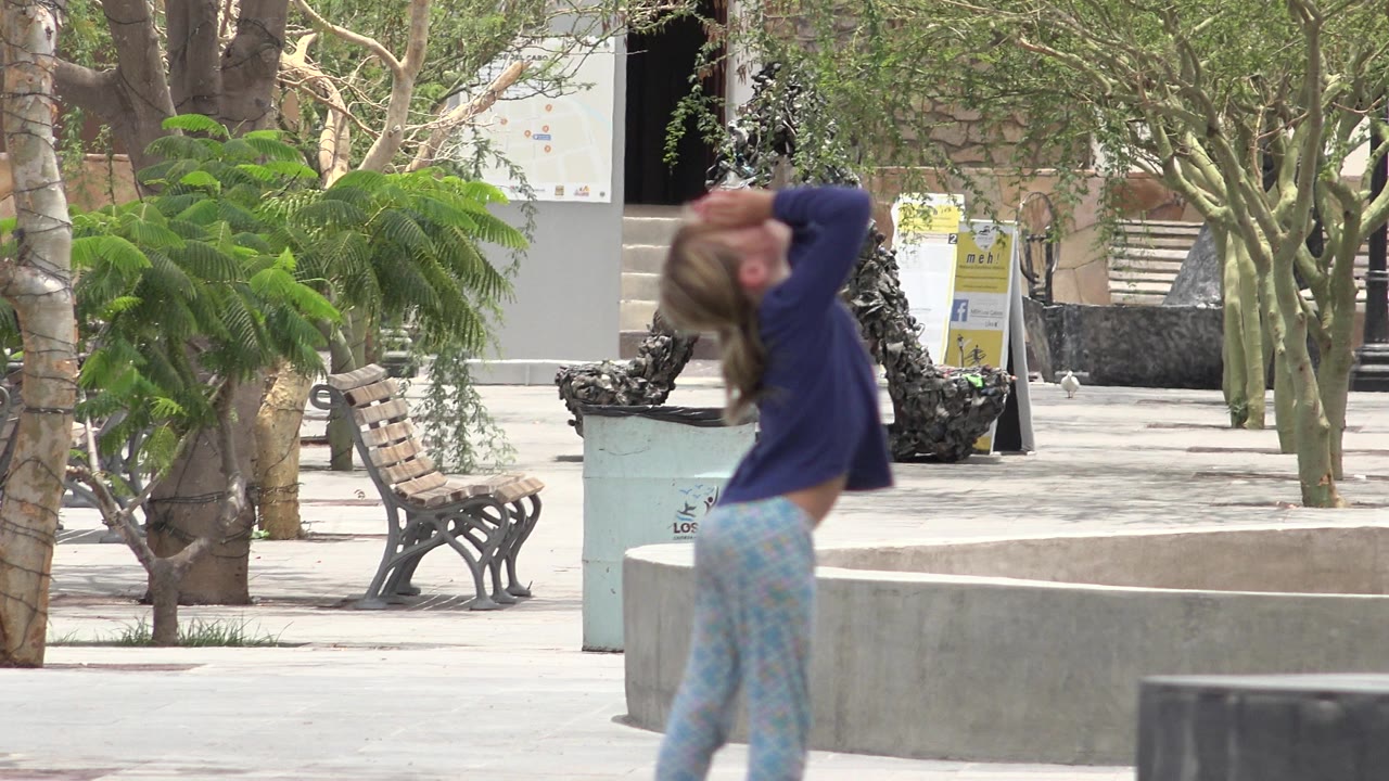 The girl remains in the foreground of the Los Cabos plaza with her arms lifted toward her eyes, with a bench, trees, and a planter in the background.
