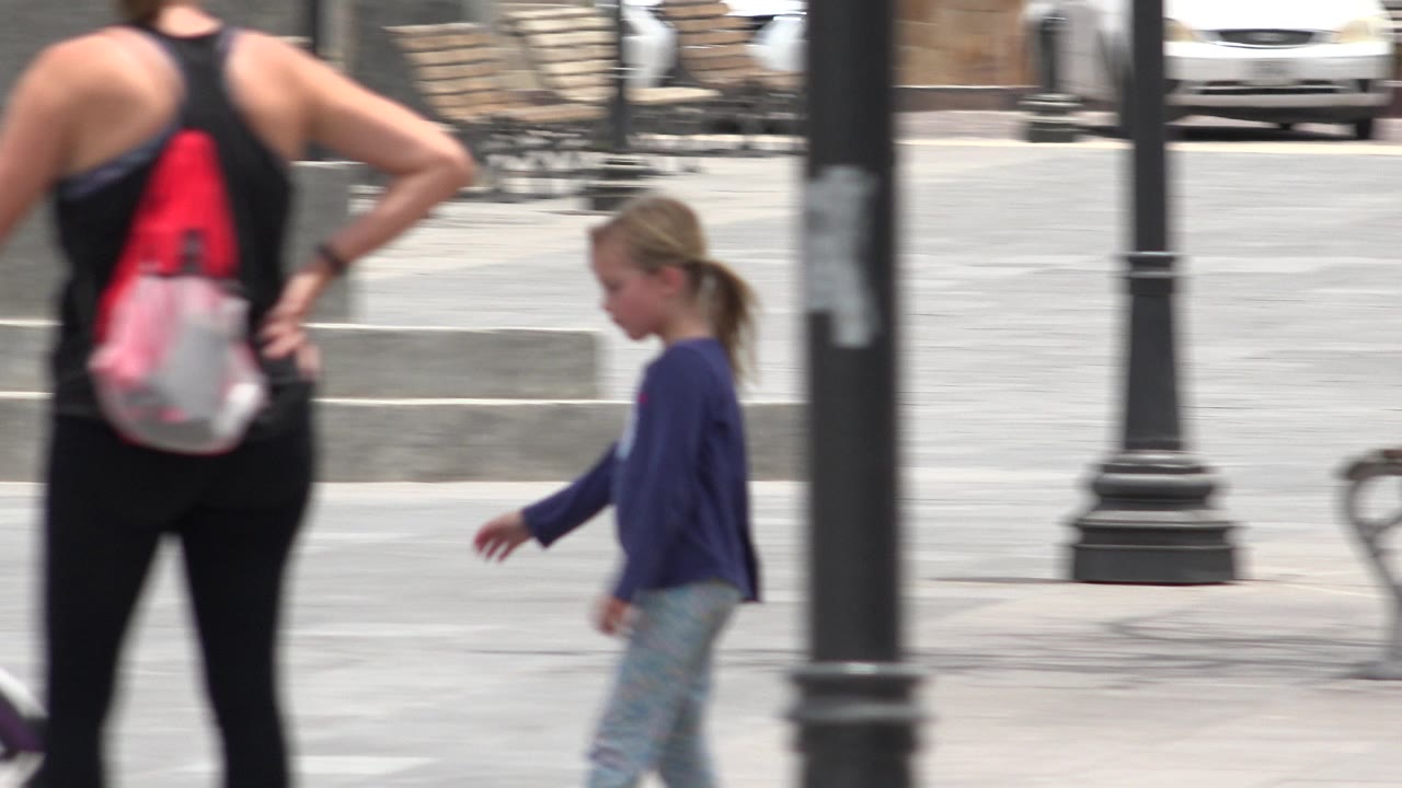 The Los Cabos square remains framed by a black lamp post as the woman with a red and white backpack stands in the foreground and the young girl walks at midframe.