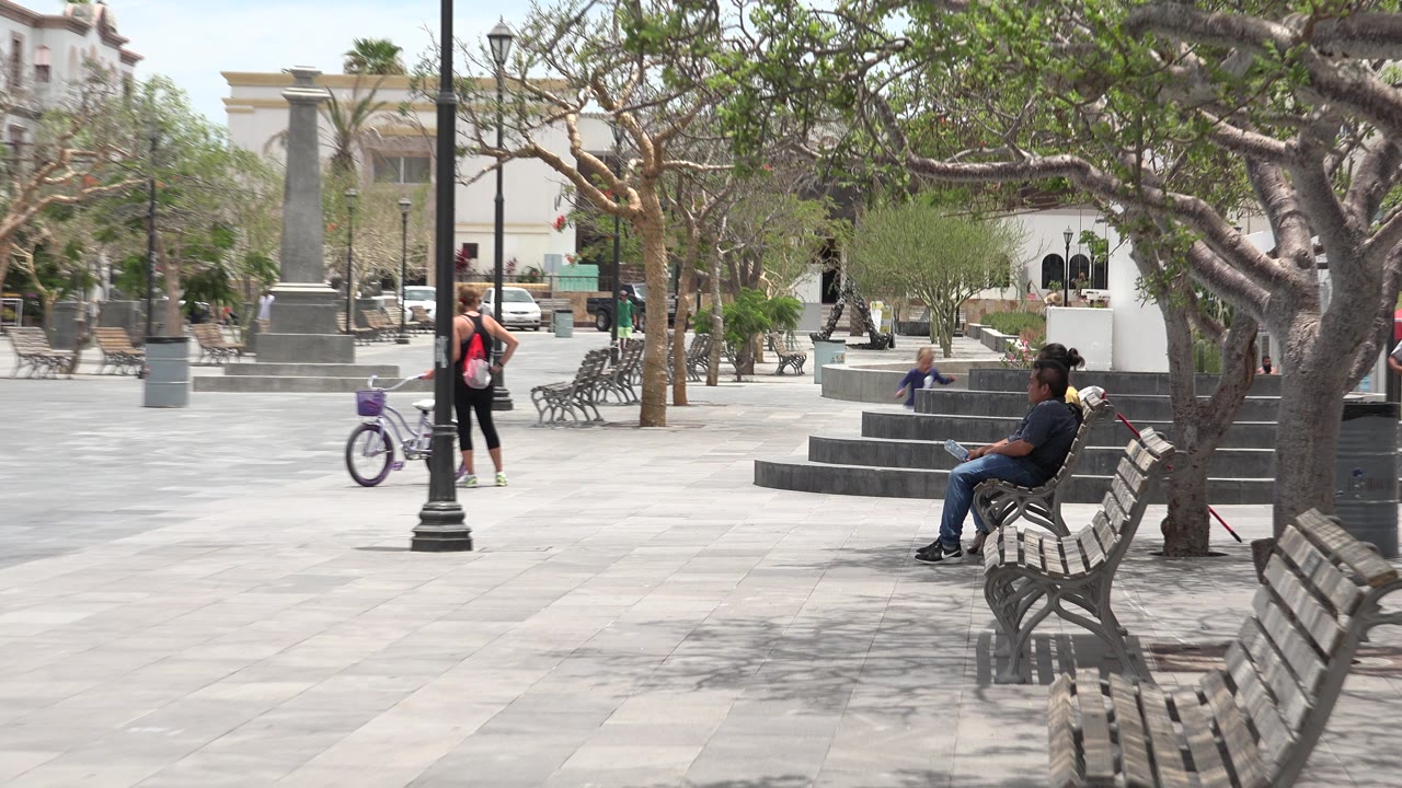 Paved public square with benches, trees, lampposts, and several people in the distance.