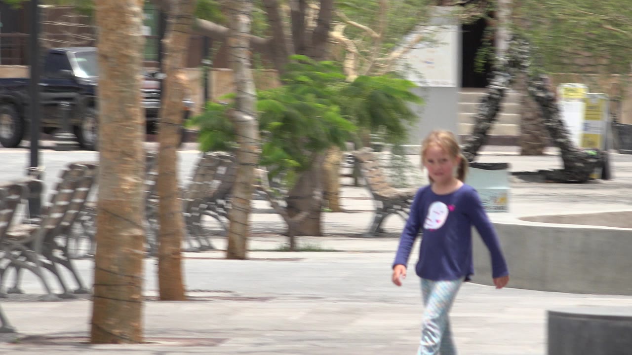 A blonde girl with a ponytail walks through a paved public area in Los Cabos, Mexico, with benches, palm trunks, planters, and parked vehicles in the background.