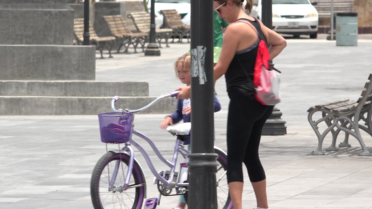 In Los Cabos, Mexico, a woman in black stands beside a young girl partly hidden by a lamppost as the girl holds a purple and white bicycle in a bright paved plaza.