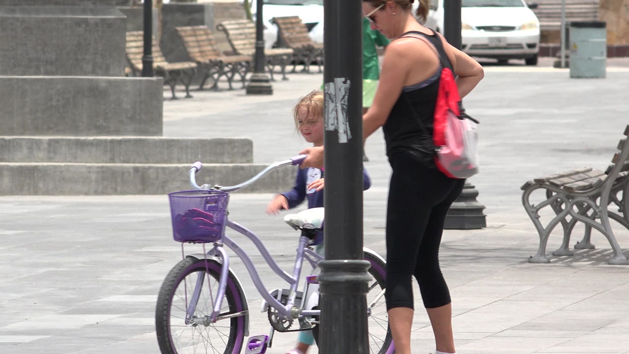 In Los Cabos, Mexico, the child reaches toward the bicycle handlebar near the center lamppost while the woman with a red backpack stands close on the right.