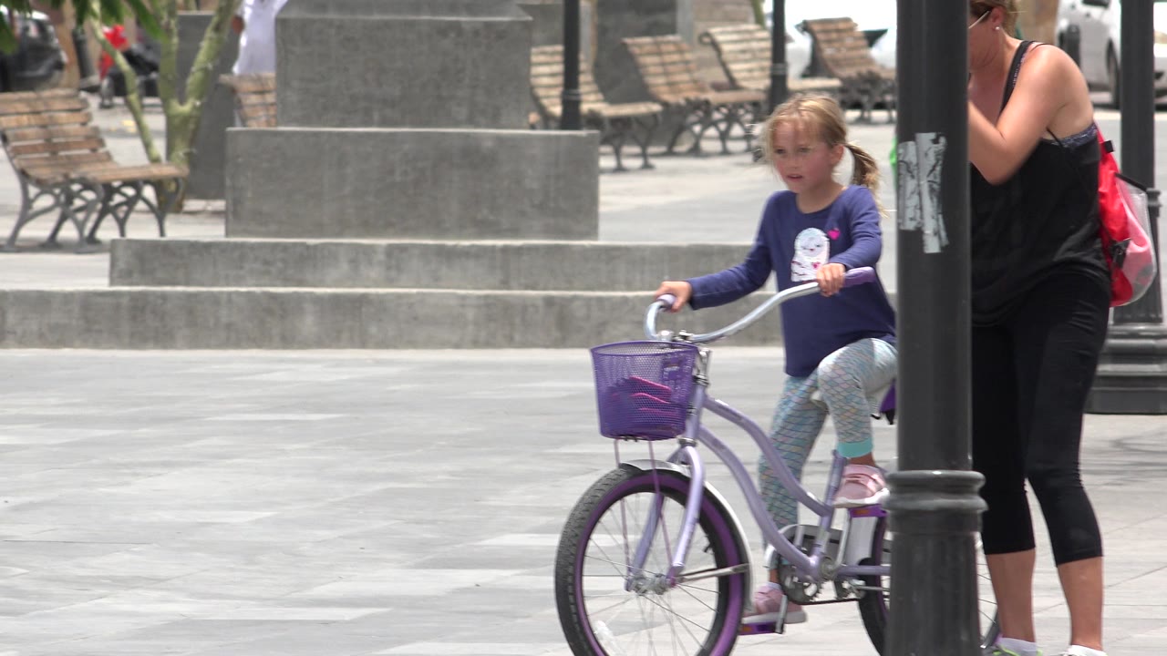 Young girl on a purple bicycle beside an adult woman in a paved plaza.