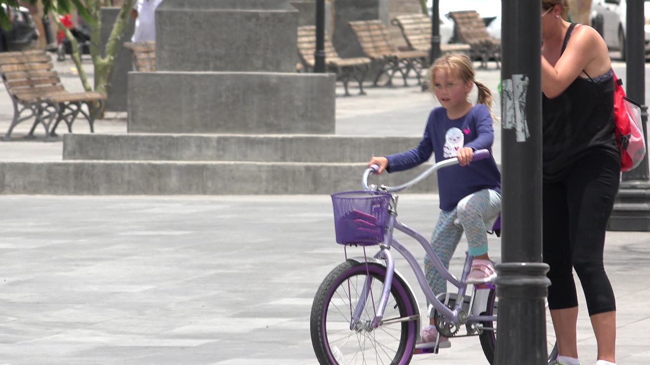 A second into the Los Cabos, Mexico scene, the girl continues forward on the purple bicycle while the adult remains beside her near a foreground lamppost.