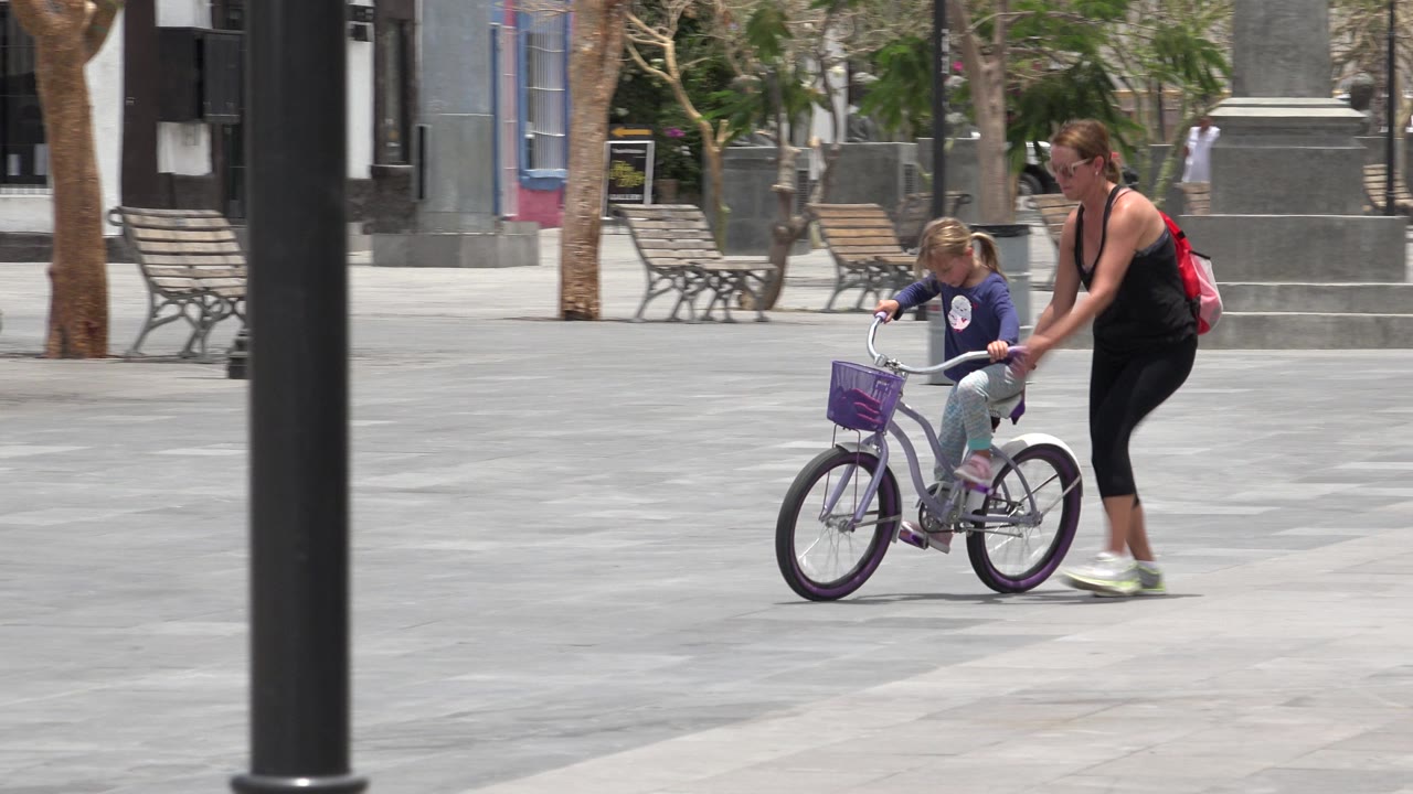 In Los Cabos, Mexico, a woman walks beside a young girl on a purple bicycle as they move across a gray paved plaza with benches and trees behind them.