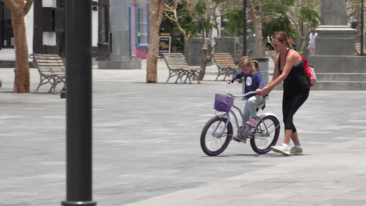 In Los Cabos, Mexico, the child rides the small purple bicycle while the woman stays close at her side, with empty benches and trees in the background.