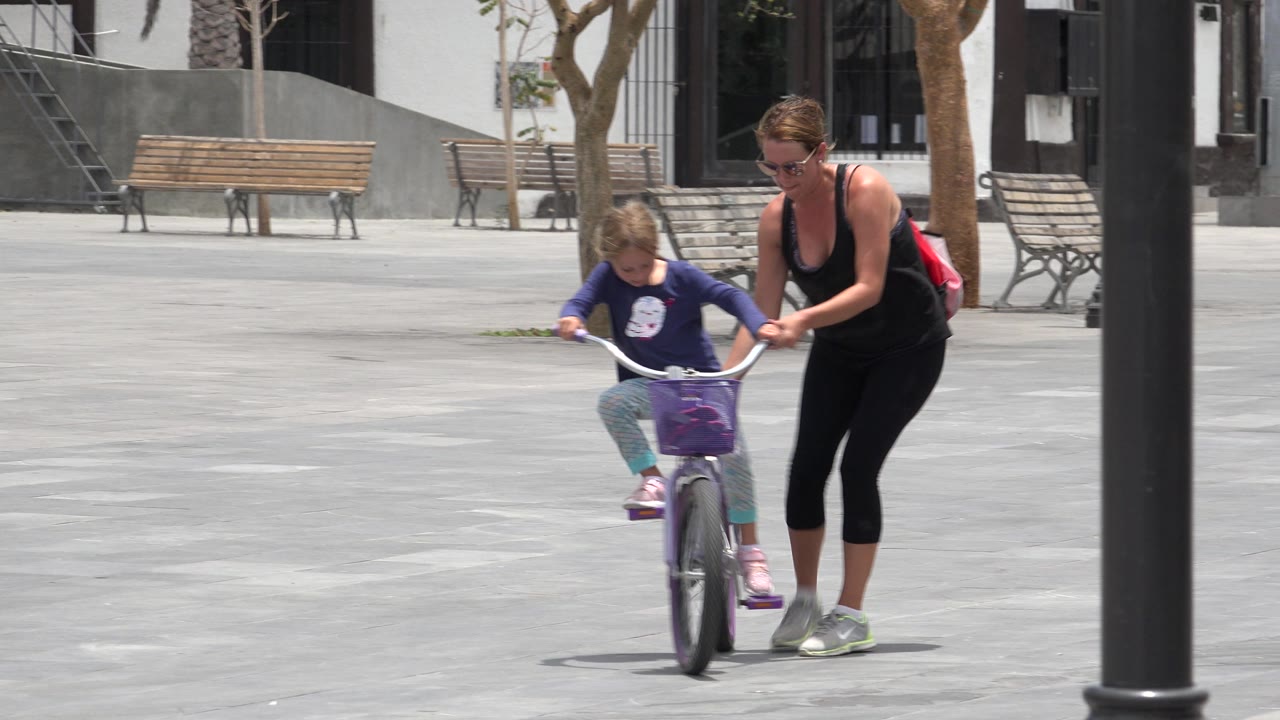Adult woman steadies a young girl on a small bicycle in a paved plaza.
