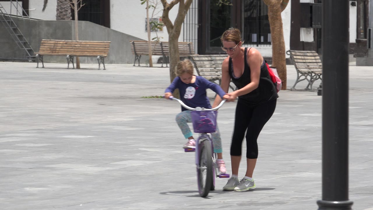 The child rides forward on the bicycle as the adult woman stays at her side, with benches and a building visible behind them in Los Cabos, Mexico.