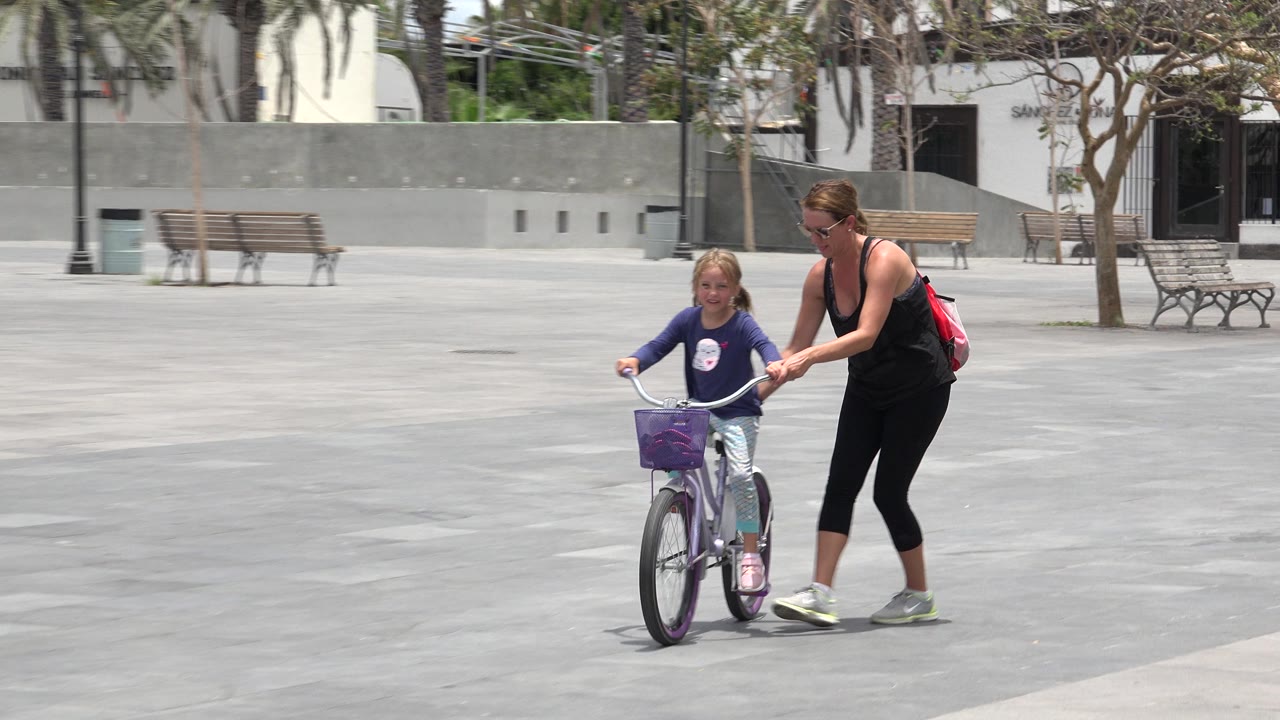 Mother walks beside a young child on a bicycle in a paved plaza.