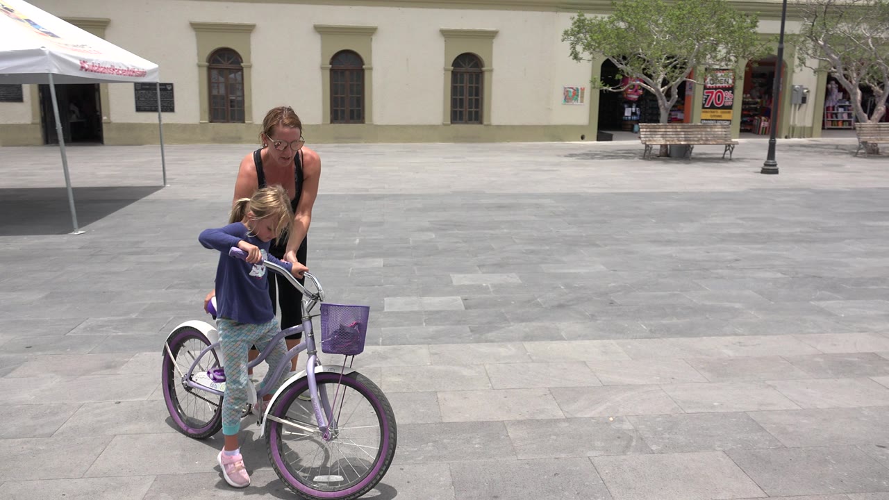 Woman helping a child balance on a purple bicycle in a paved plaza.