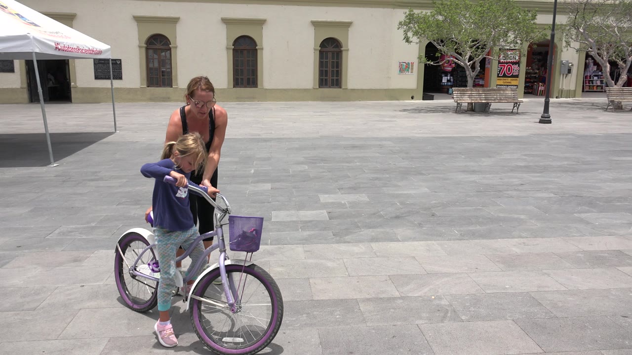 In Los Cabos, Mexico, the child stays over the purple bicycle while the woman stands directly behind, with open paving and a pale building across the background.