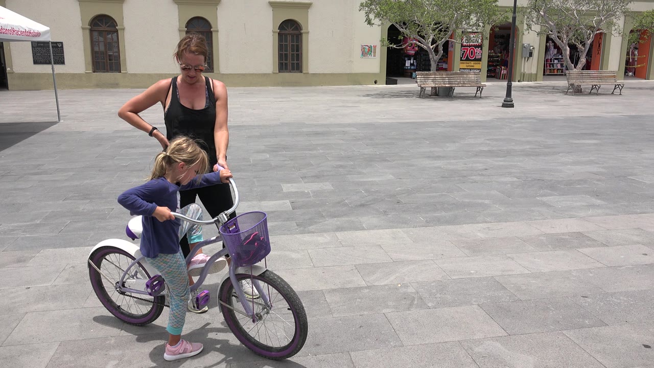 In Los Cabos, Mexico, a mother stands close behind a young girl straddling a white and purple bicycle at the start of a riding lesson in a paved plaza.
