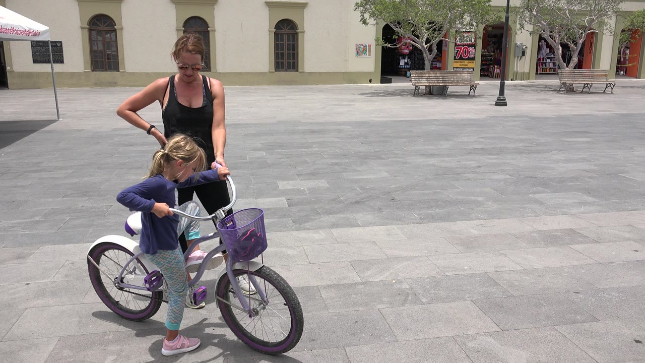 In Los Cabos, Mexico, the girl leans over the handlebars of the white bicycle as her mother stands behind her on the open gray plaza paving.