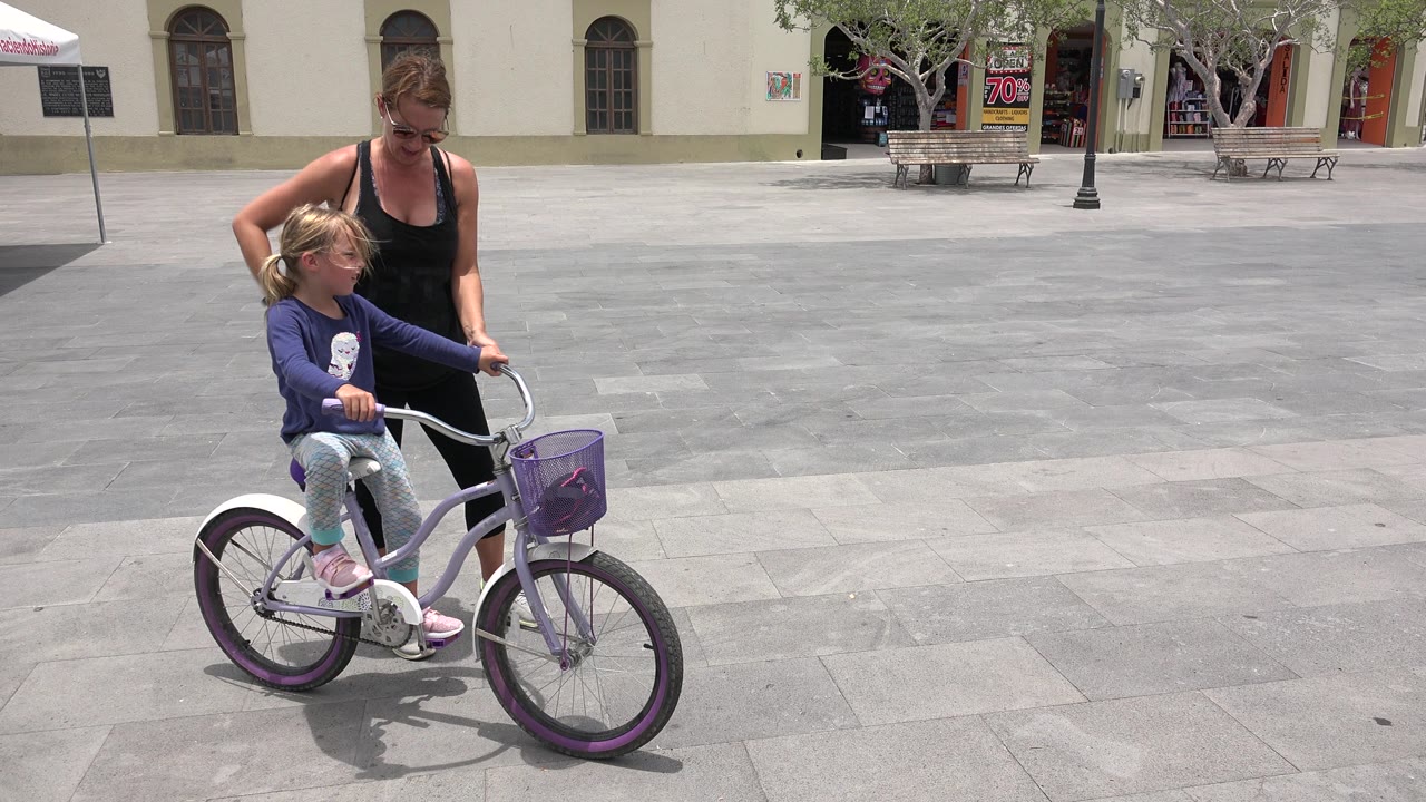 Woman walking beside a young girl on a purple bicycle in a paved plaza.