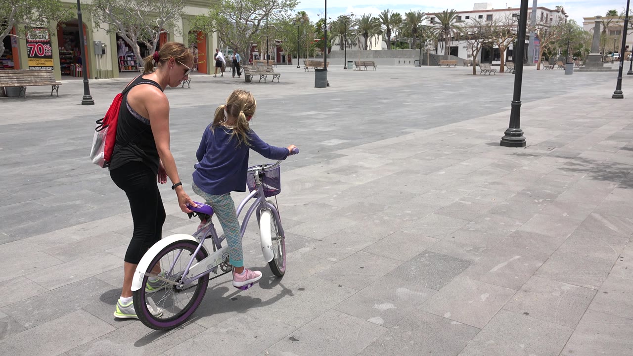 Woman guiding a young girl on a bicycle in an open plaza.