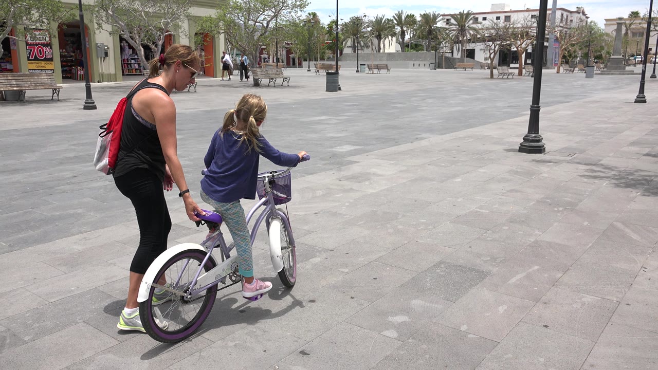 The woman and young girl continue across the Los Cabos plaza, with the bicycle angled forward and benches, trees, and a storefront visible behind them.