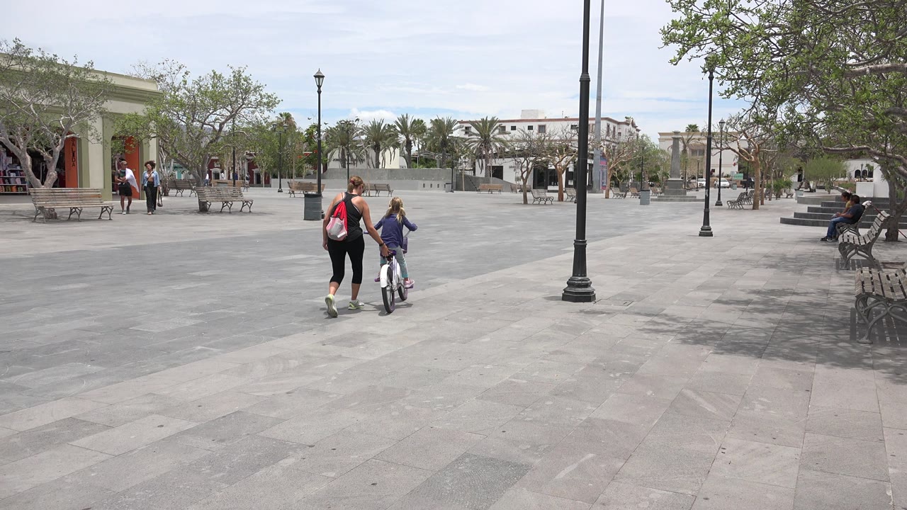 In Los Cabos, Mexico, an adult in dark clothes walks beside a child on a small bicycle across a wide gray courtyard with benches, trees, and lamp posts.