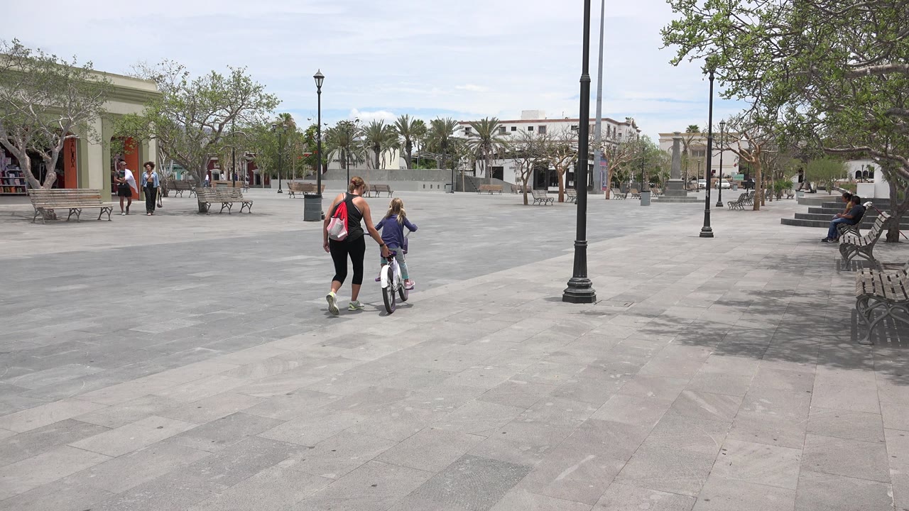The child on the small bicycle moves farther into the Los Cabos courtyard while the adult remains close, with benches and a large tree visible to the right.