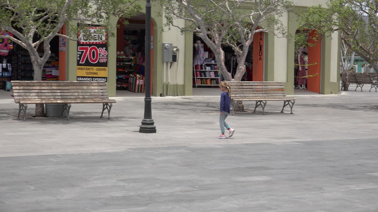 In Los Cabos, Mexico, a blonde girl walks across a wide paved plaza with wooden benches, bare-branched trees, a lamp post, and open shopfronts behind her.