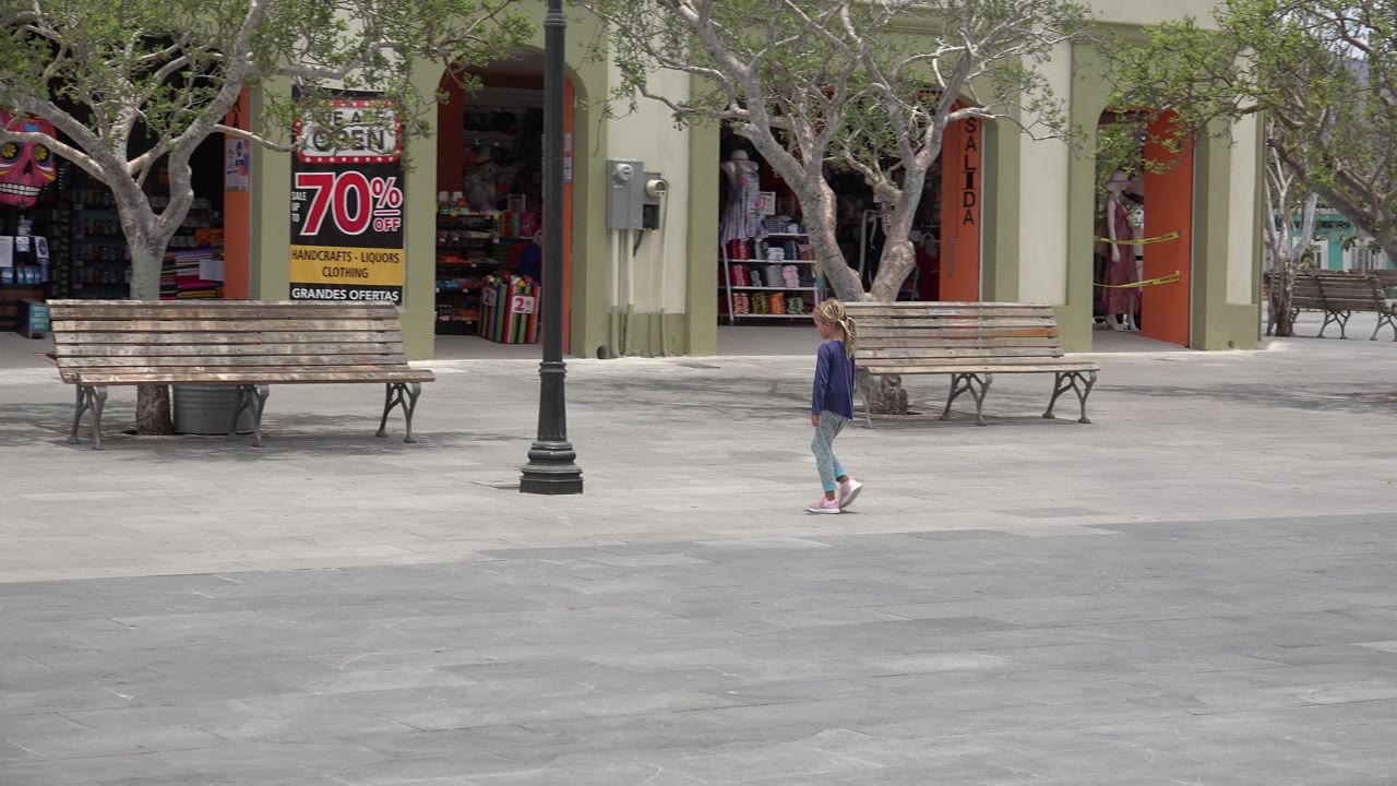 The girl continues across the Los Cabos plaza, passing in front of wooden benches and trees while shop interiors and a large discount sign remain in the background.