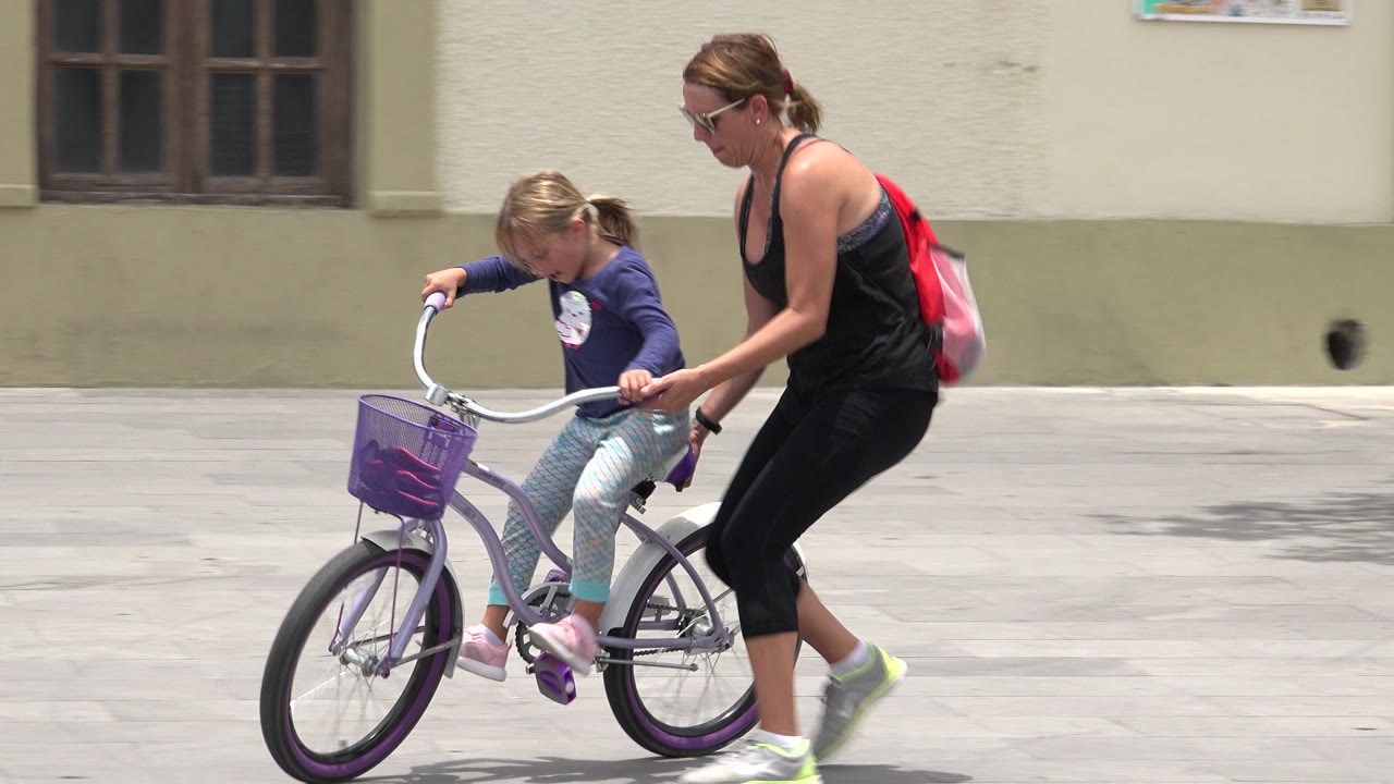 Mother steadies a child on a small purple bicycle.