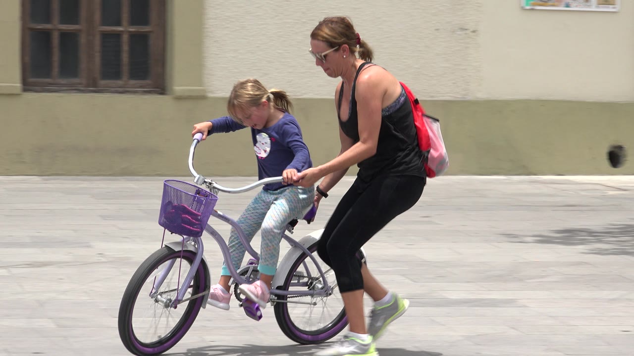 In Los Cabos, Mexico, the girl keeps both hands on the handlebars while the woman stays close behind the bike on the open paved area.