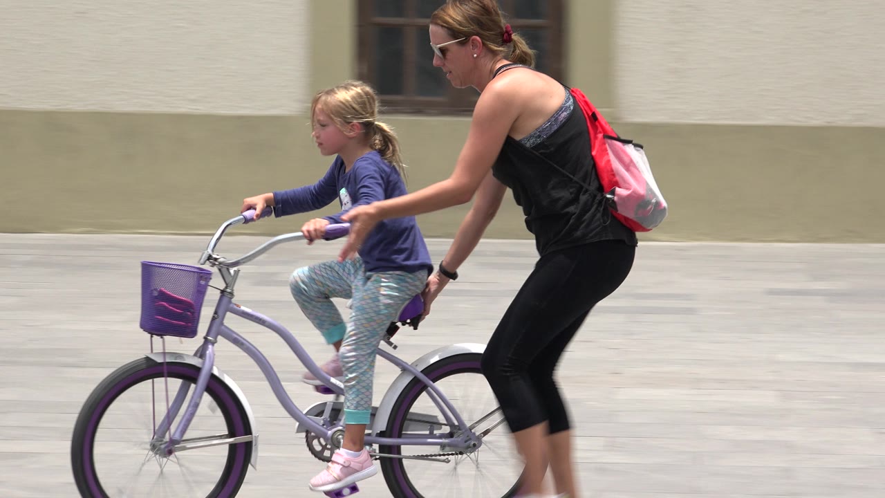 In Los Cabos, Mexico, an adult woman walks beside a young child riding a small purple bicycle across pale paving in bright daylight.