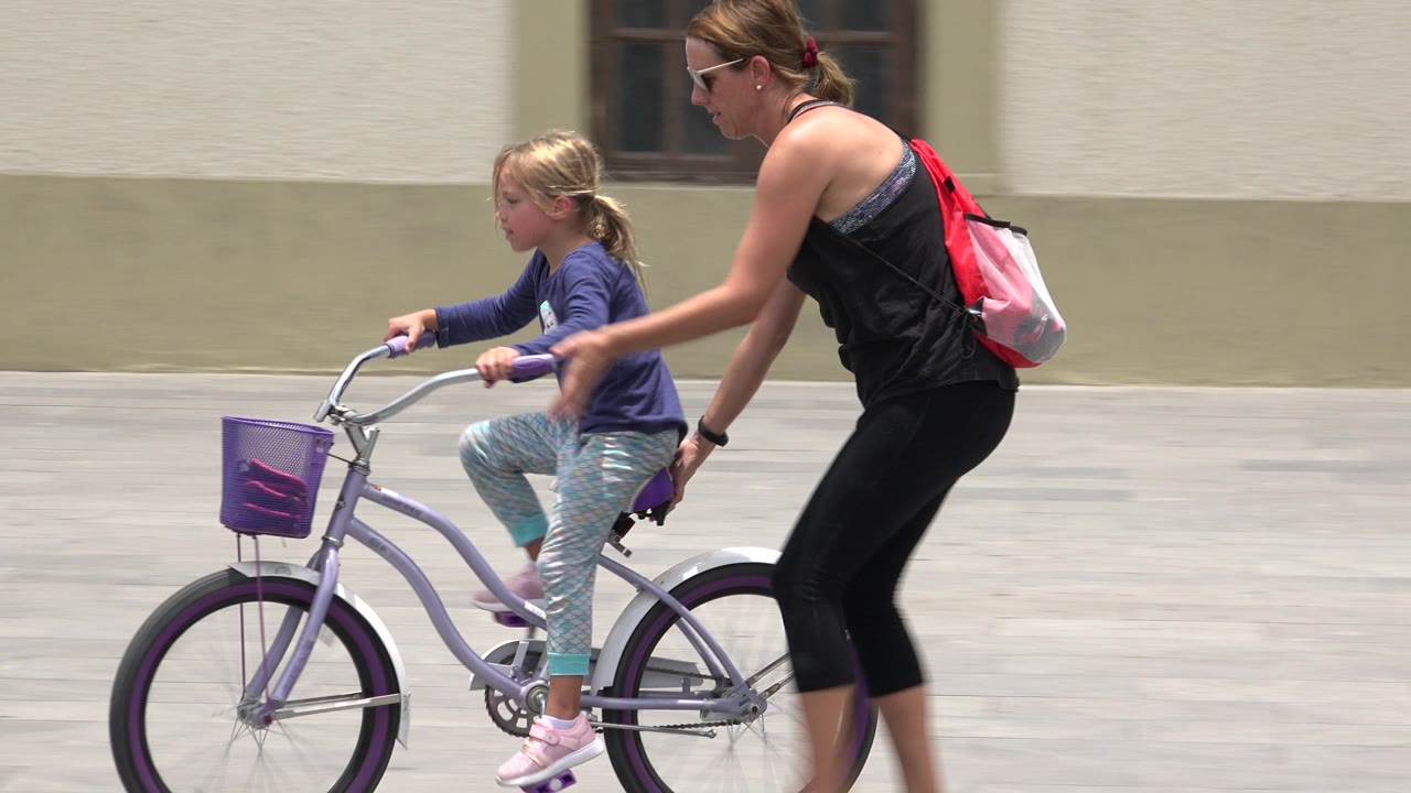 The child pedals the purple bicycle in Los Cabos, Mexico, while the woman leans forward with one hand near the back of the seat.